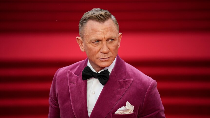 A man wearing a velvet suit jacket and black bowtie poses with stern facial expression on a red carpet