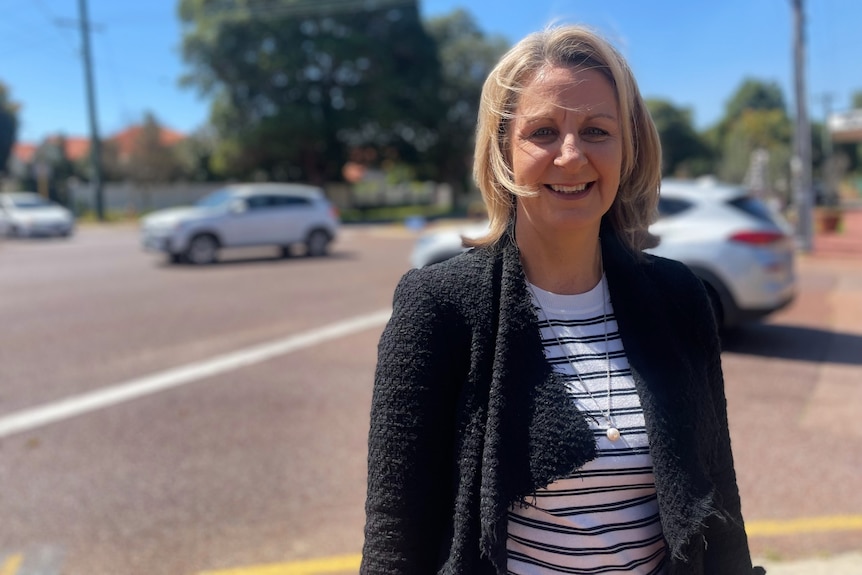 A woman stands outside with a road in the background. There are cars on the road.