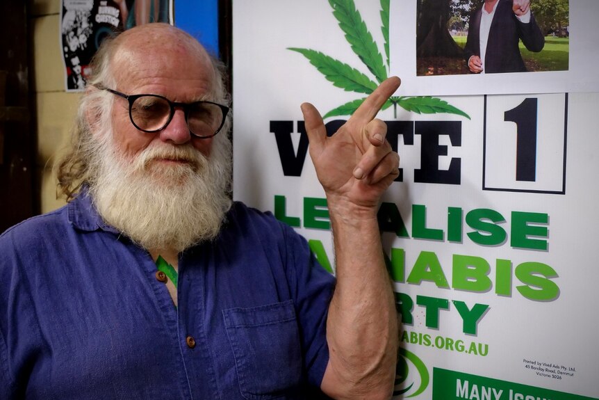 A man with black glasses and white hair and beard pointing to a vote sign with a cannabis leaf