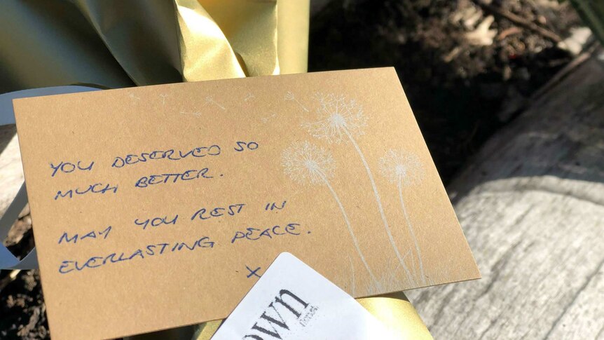 A note on a bouquet of flowers on a log reading "You deserved so much better. May you rest in everlasting peace."