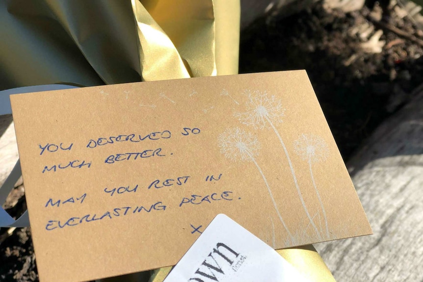 A note on a bouquet of flowers on a log reading "You deserved so much better. May you rest in everlasting peace."