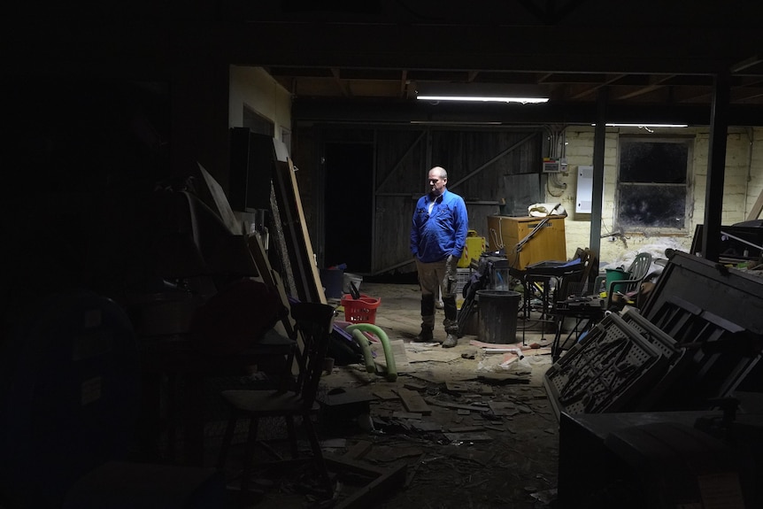 A wide shot of a man standing in a crowded garage at night time. A small amount of light illuminates him