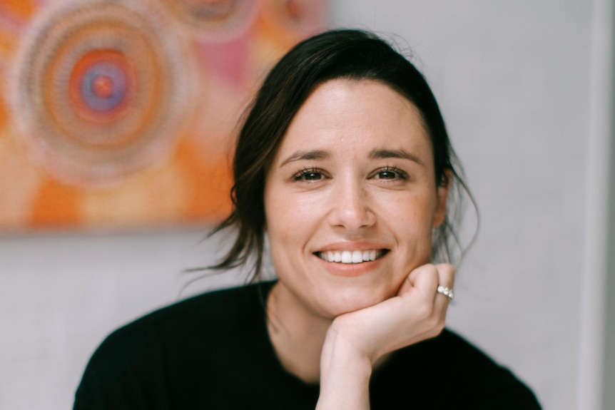 a woman with dark hair smiles directly at the camera leaning her chin on her hand