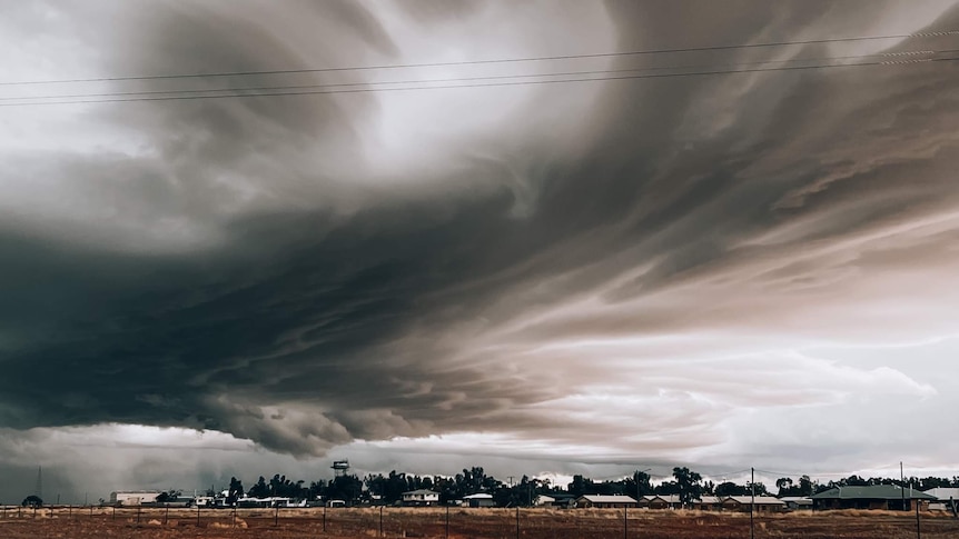 a big black and grey cloud is swirling over a small outback town with houses and red dirt