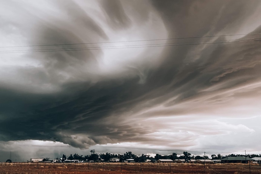 a big black and grey cloud is swirling over a small outback town with houses and red dirt
