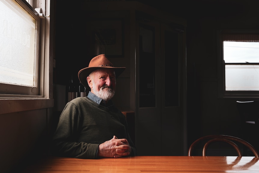 A man in an Akubra hat sits at a table by a pub window smiling.