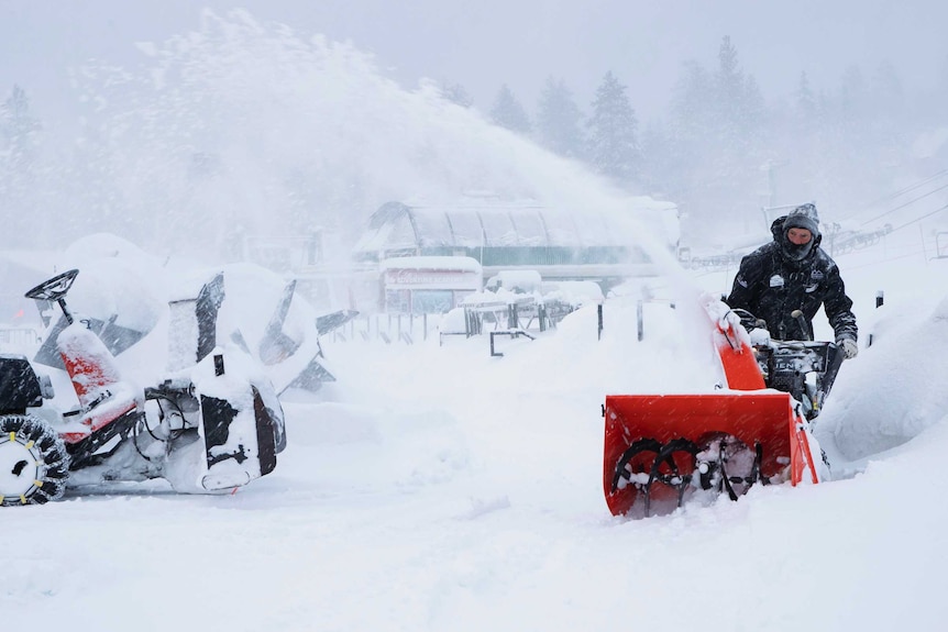 A man on a motorised device moves snow from an accessway. Heavy snow can be seen on buildings all around.