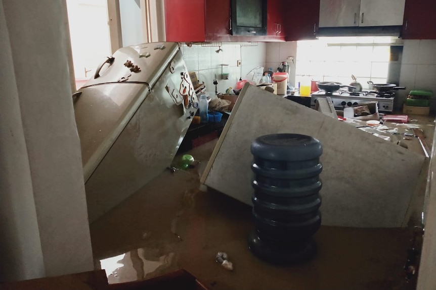 A fridge and other kitchen items floating on their side in muddy brown water flooding a kitchen.