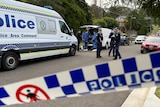 Police tape across a street with a number of police cars and people in forensic suits standing next to police vehicles.
