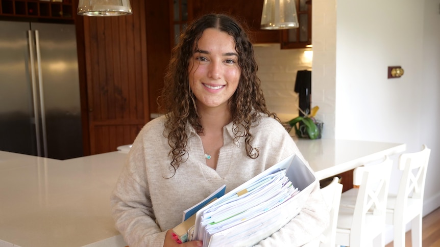 Sheridan wears a white jumper and jeans and holds a large binder while sitting down in her kitchen.