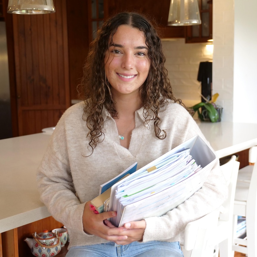 Sheridan wears a white jumper and jeans and holds a large binder while sitting down in her kitchen.