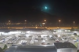 A security camera at an airport captures a car park and stationary airplanes. Behind them a meteor is falling. 