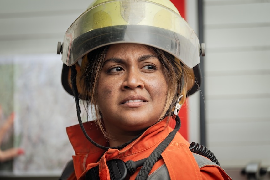Close up of Jessica Mauboy's concerned, slightly ashed face, sandwhiched between a firefighter helment and suit. 