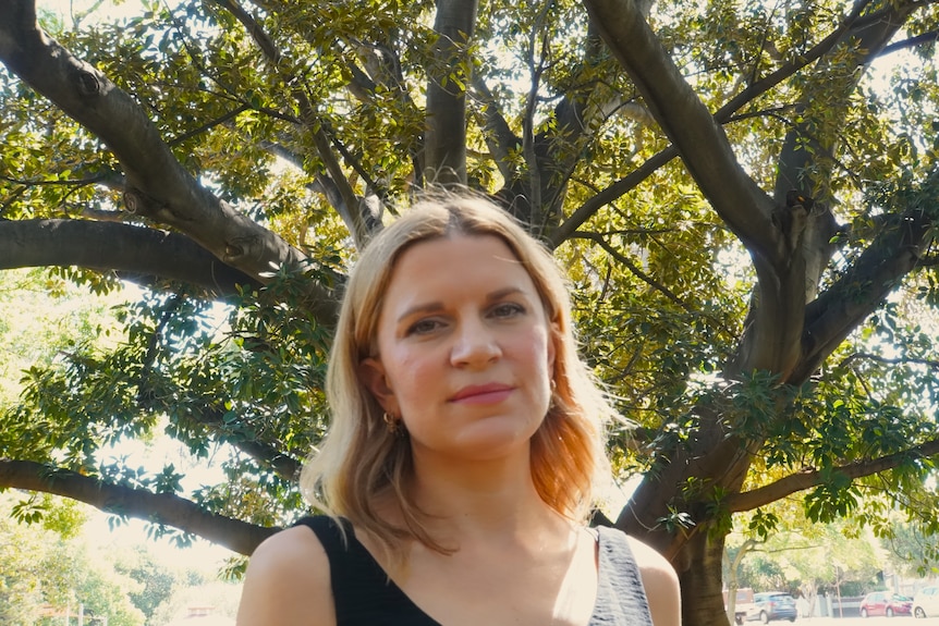 Woman looks at camera and stands in a park with large tree behind her