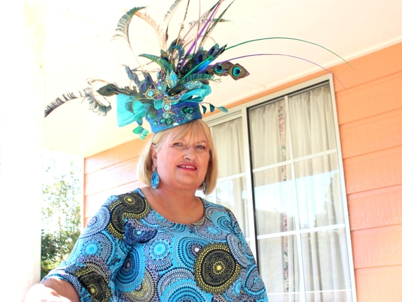 Caren Stevenson stands with large blue hat creation with feathers on her head