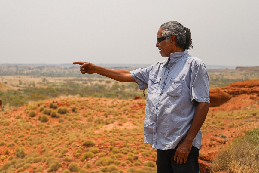 An Indigenous man wearing a light blue shirt, standing in red-sand country, pointing.