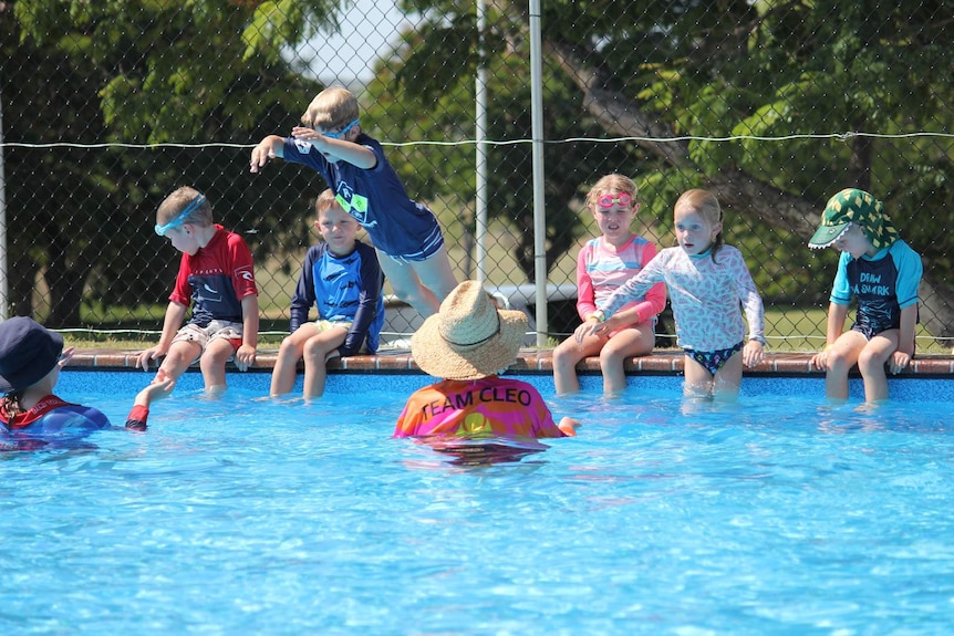 Children sit on the side of a swimming pool and jump in as instructed by a teacher