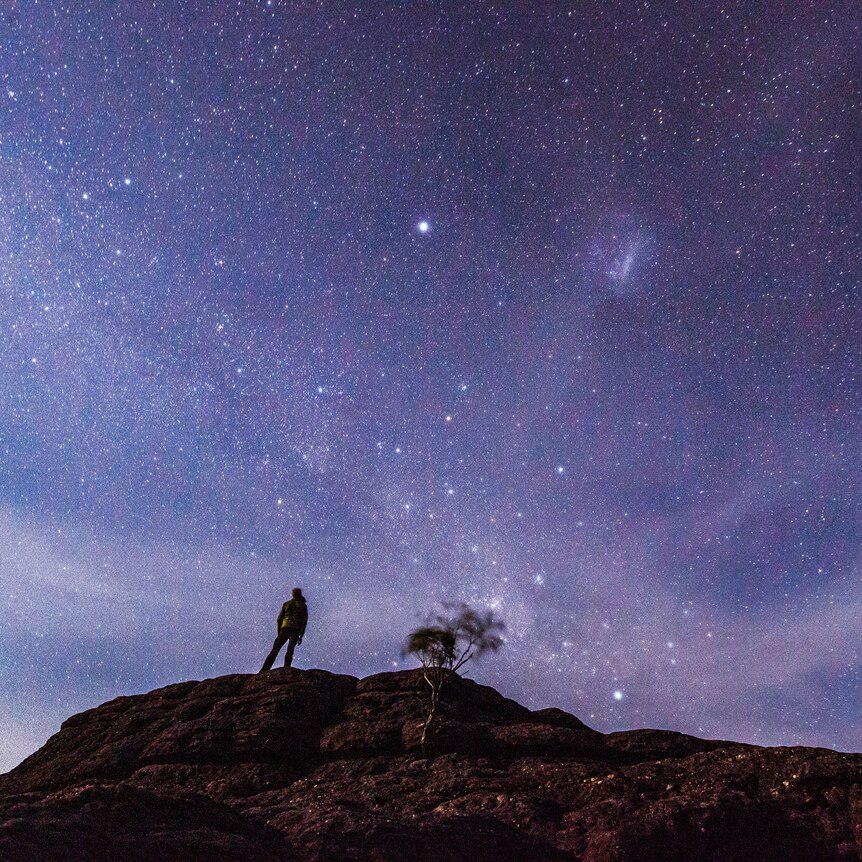 Image of someone standing on a rock formation looking up at the stars in the night sky. 