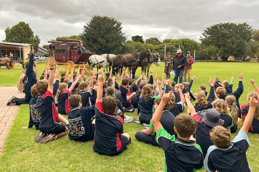 primary school students raise their hands to ask questions of a stagecoach driver with five horses