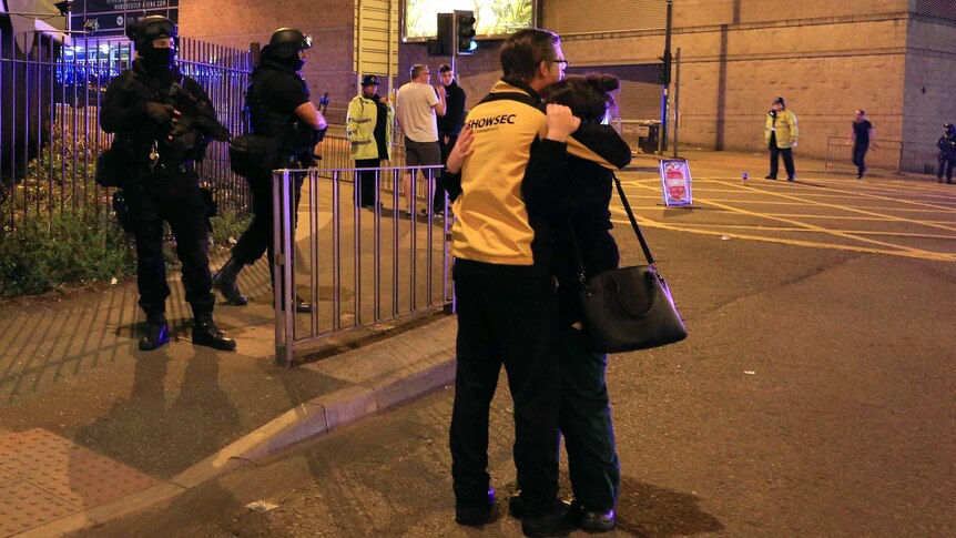 Two people hug outside Manchester Arena while armed police stand guard.