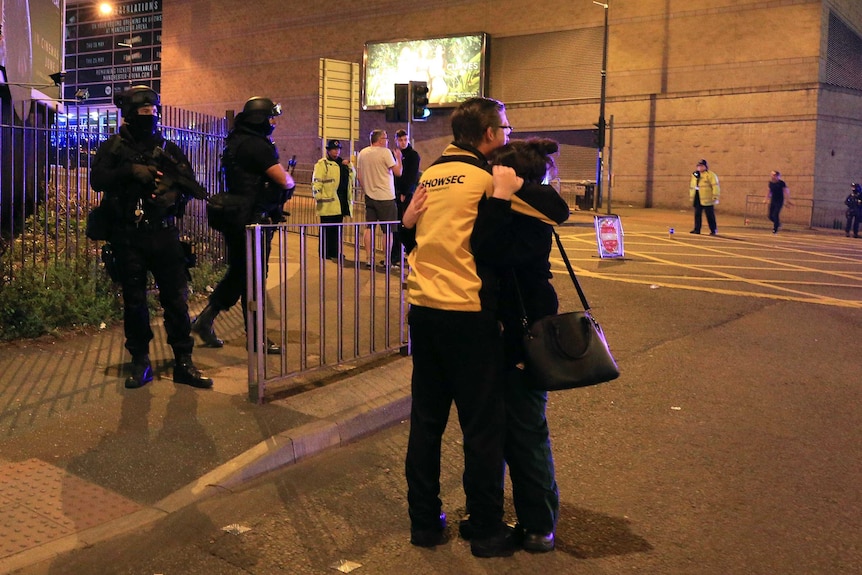 Two people hug outside Manchester Arena while armed police stand guard.