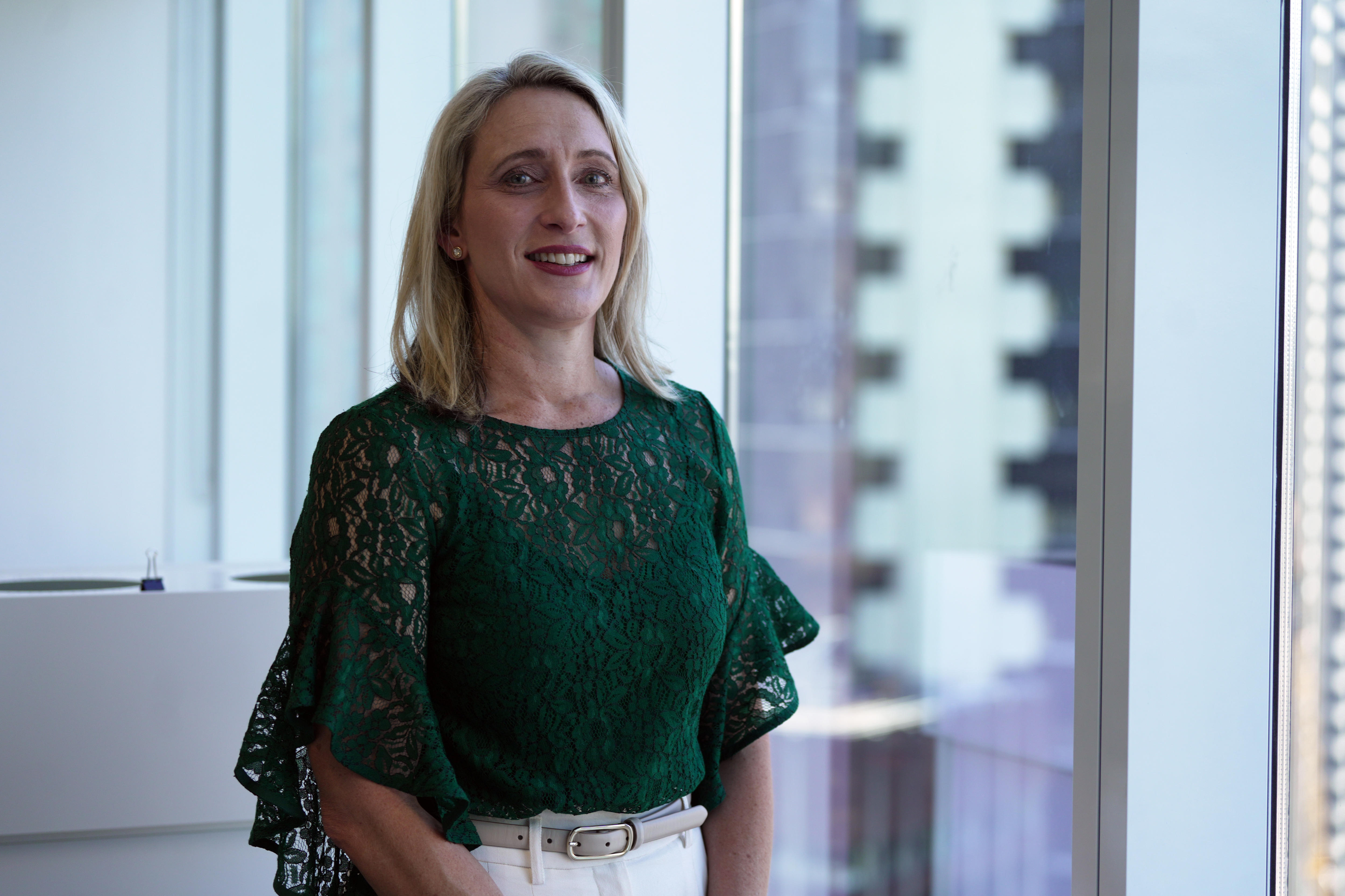 Kylie Rendina, a blonde woman wearing a bottle green shirt, stands in front of an office window.