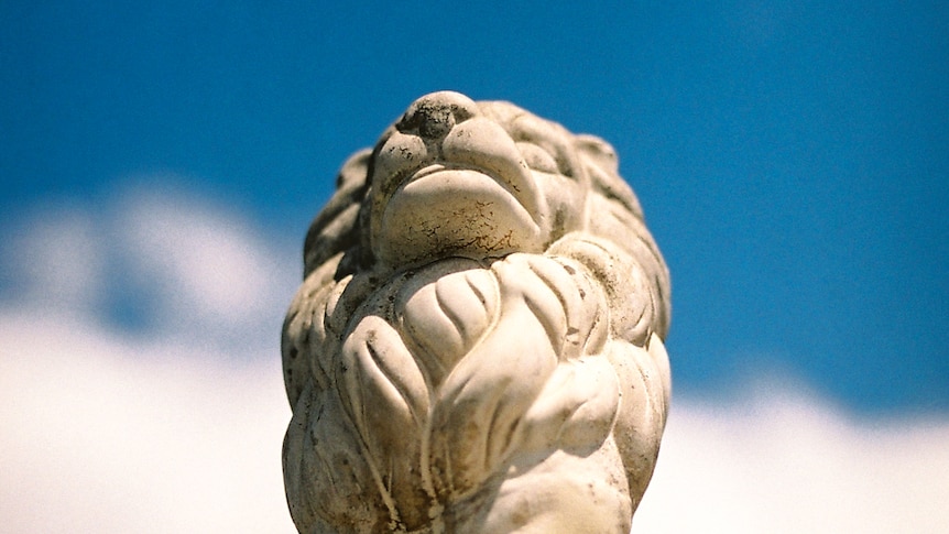 You look up at a decaying white stone lion at a blue sky.