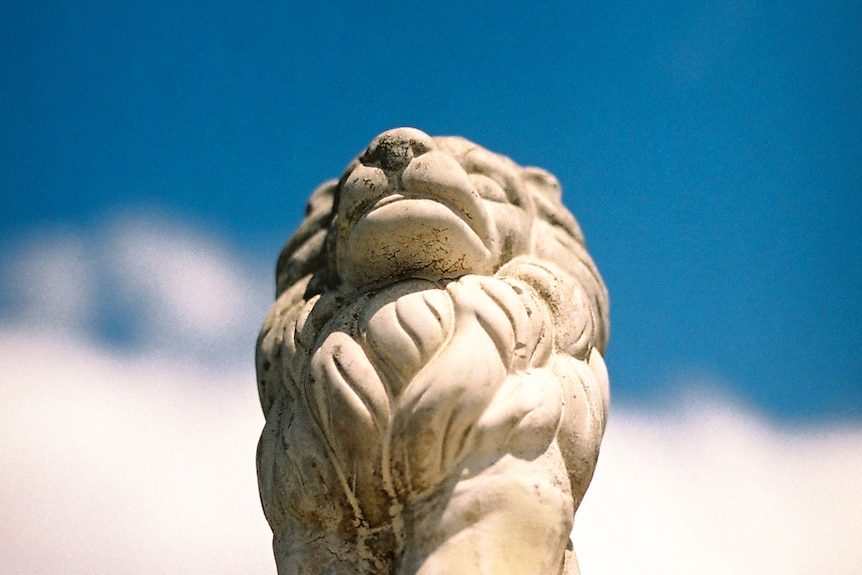 You look up at a decaying white stone lion at a blue sky.