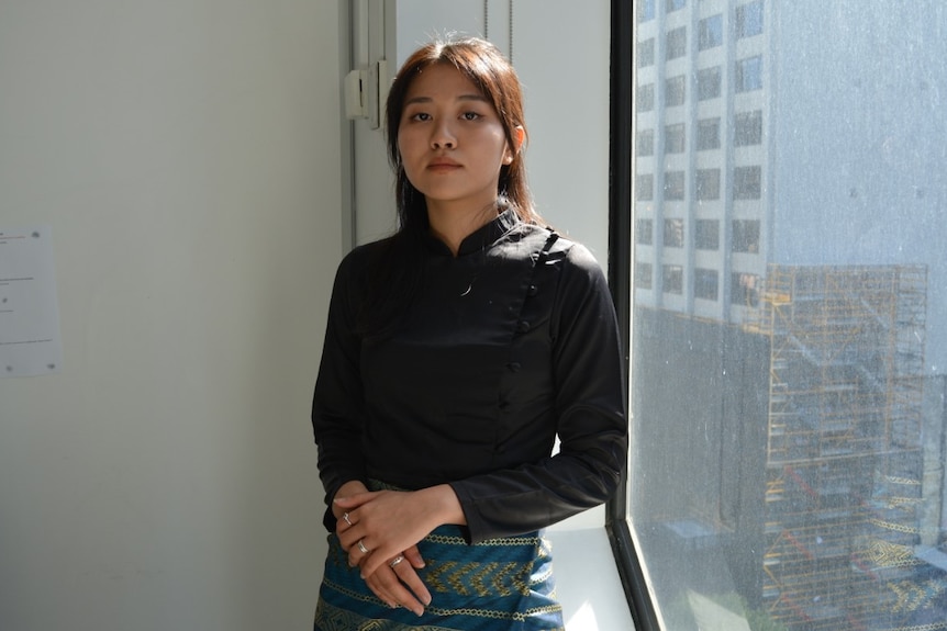 Kyi Phyu Moe Htet in a blue traditional Burmese skirt stands unsmiling in front of a window.