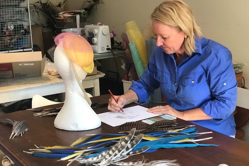 Felicity Brown sits at a table sketching, with a hat form and several feathers in front of her.