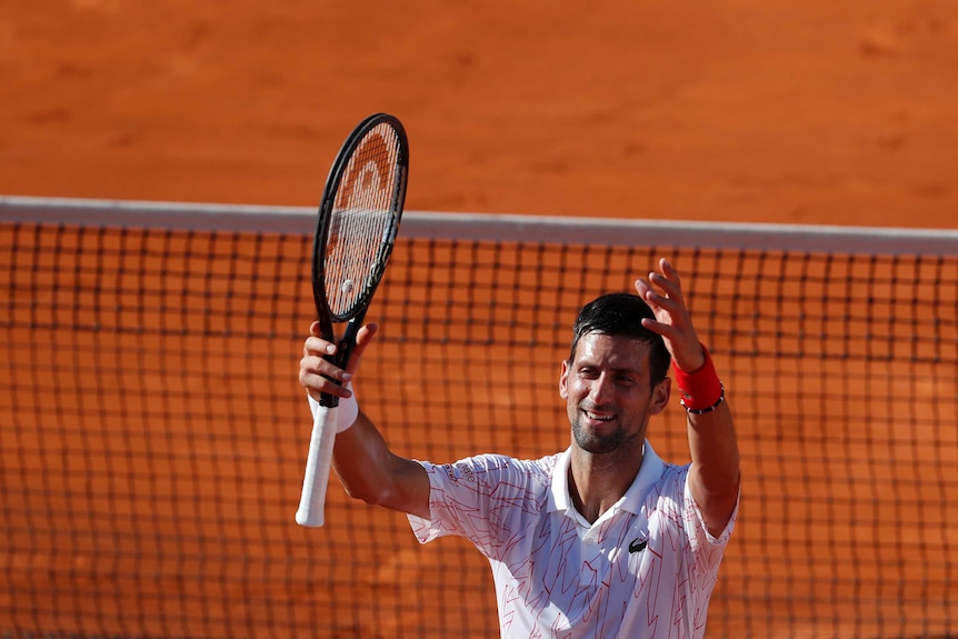 A tennis player stands in front of the net, with his arms raised in triumph and salute to the crowd.