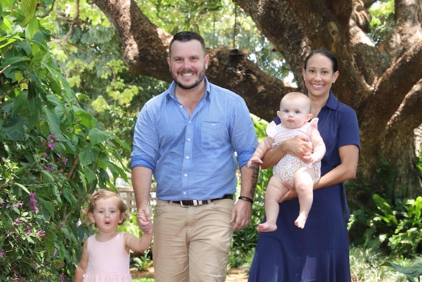 Phillip and Jenna Thompson walk through a green park with trees in the background Jenna holds baby small child holds Phil's ha