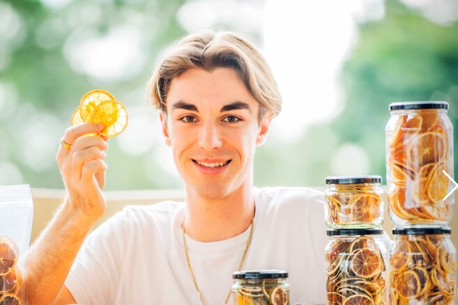 A young man holds up dried citrus slices with jars of dried fruit and fresh fruit near him.