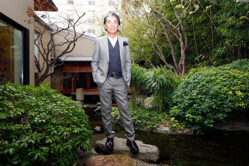 Kenzo Takada is in a silver suit standing on stones atop a ceremonial pond in the middle of a residential Japanese garden.
