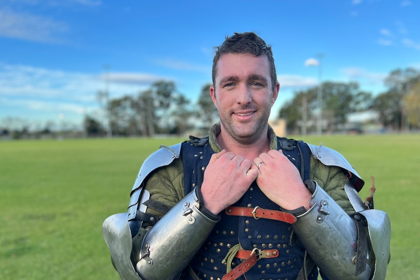 Smiling man, wears silver armour on shoulders and arms, a green long sleeve undershirt and blue chest piece, on a green field.