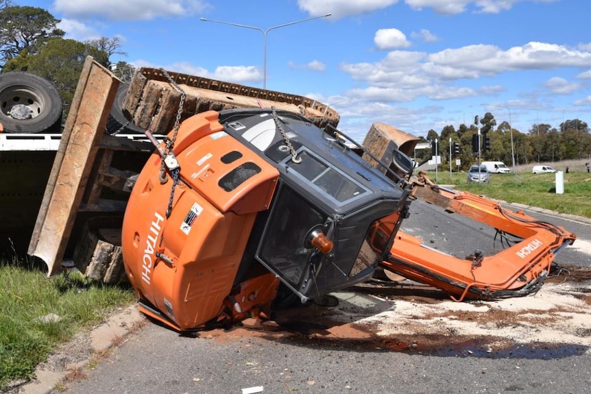 An orange excavator on its side on a road, with an upturned truck next to it.