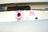 A close-up of the side of a white ship shows a Japanese flag and a white banner carrying Japanese characters.