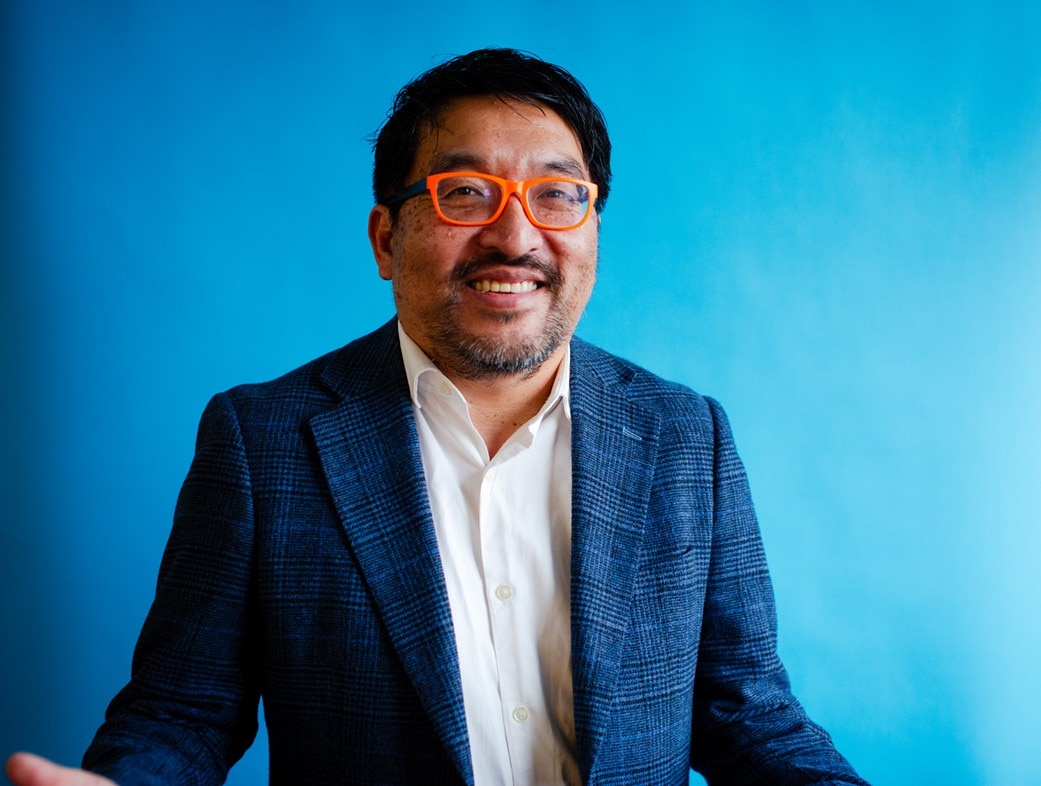 A corporate headshot of John Chan in orange glasses.
