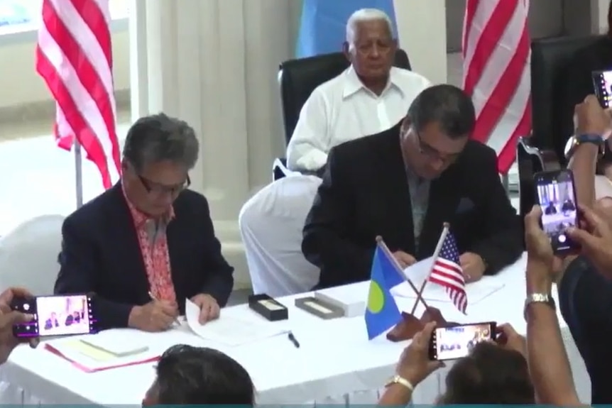 Two men seen sitting at table signing documents in front of audience.