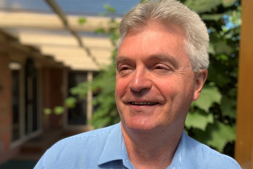 A smiling man in a blue shirt looks off to the side in a courtyard garden.