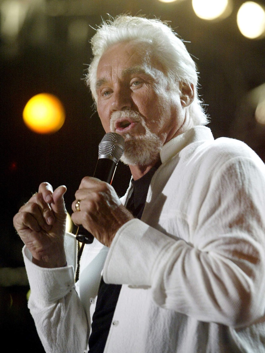 Kenny Rogers stands on stage singing into a microphone wearing a white suit