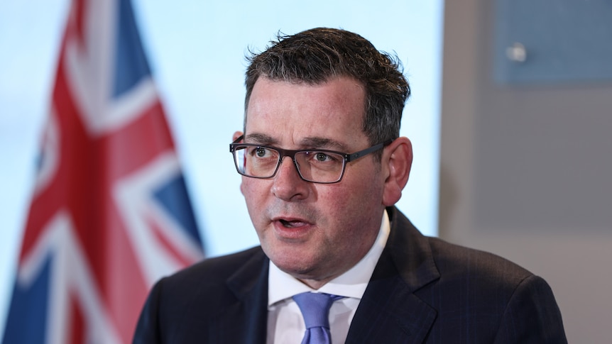 Daniel Andrews wearing glasses and black jacket with the british flag behind him