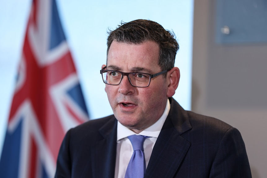 Daniel Andrews wearing glasses and black jacket with the british flag behind him