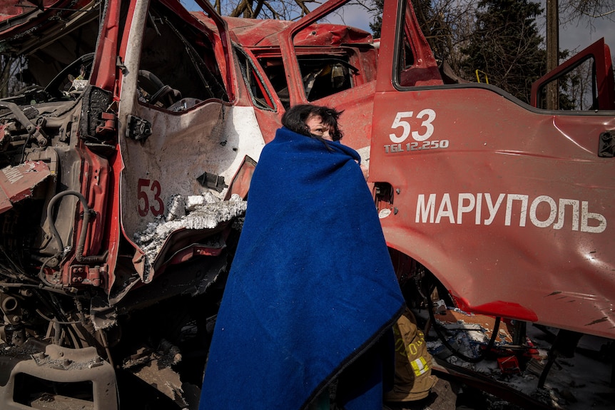 一名妇女在一辆损坏的消防车附近用蓝色毯子盖住自己。