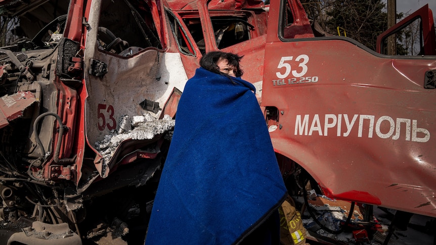 A women covers herself with a blue blanket near a damaged fire truck.