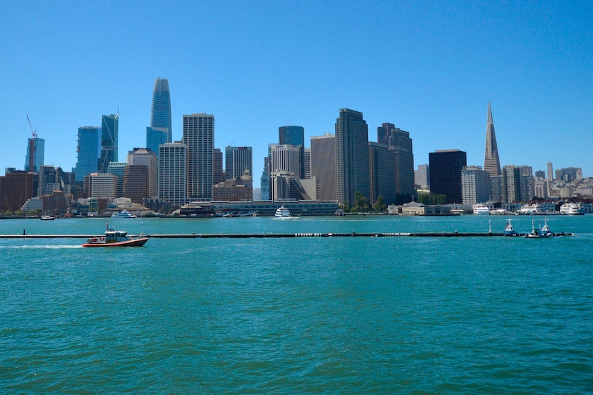 A ship tows a large buoyant trash-collecting device toward the Golden Gate Bridge in San Francisco. The city skyline is visible