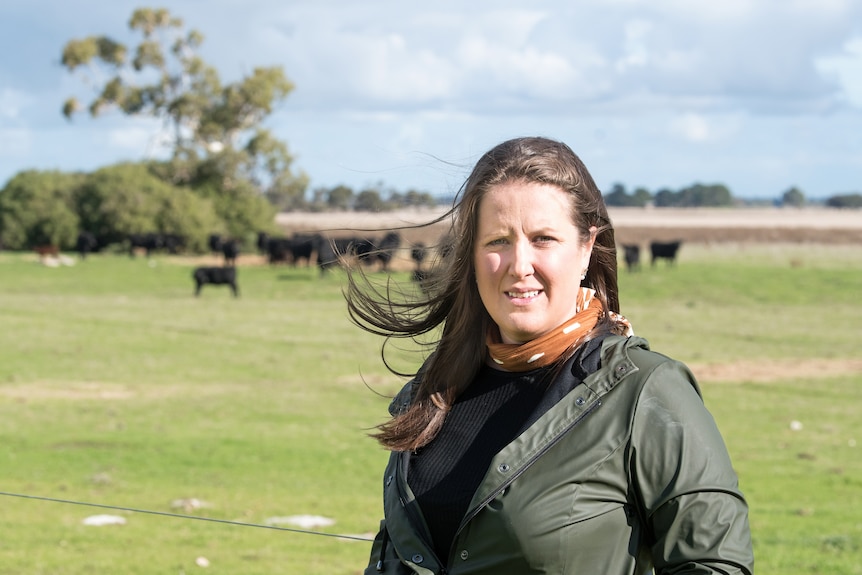 A woman in a green jacket and long brown hair stands smiling at the camera with a paddock with cows behind her