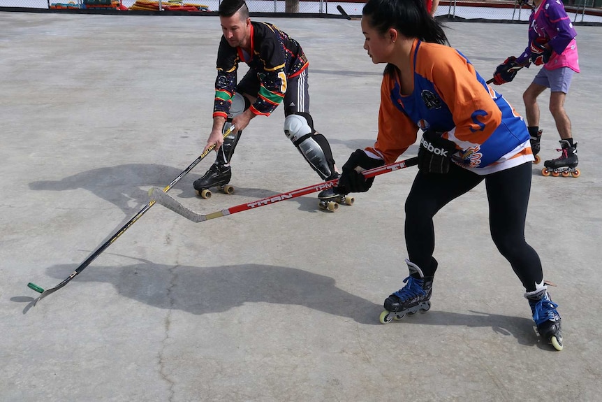 Players in brightly coloured clothing play roller hockey on a grey concrete court.