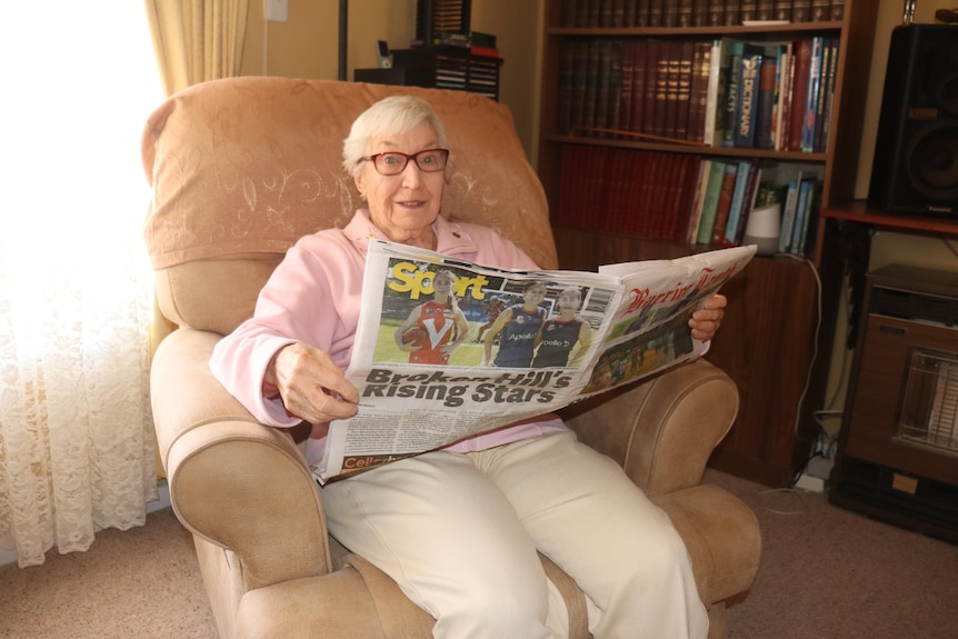 An elderly woman sitting in an armchair holding an opened newspaper smiling