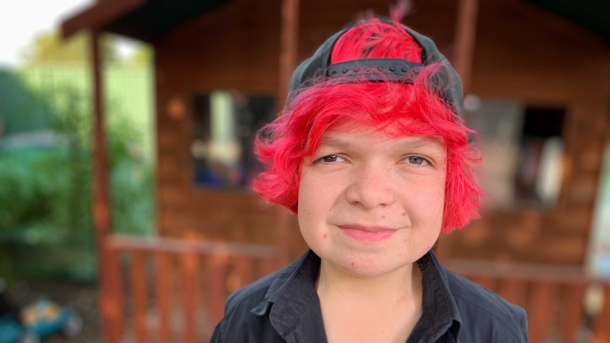 Girl with bright red hair, backwards cap and black shirt smiles at camera.